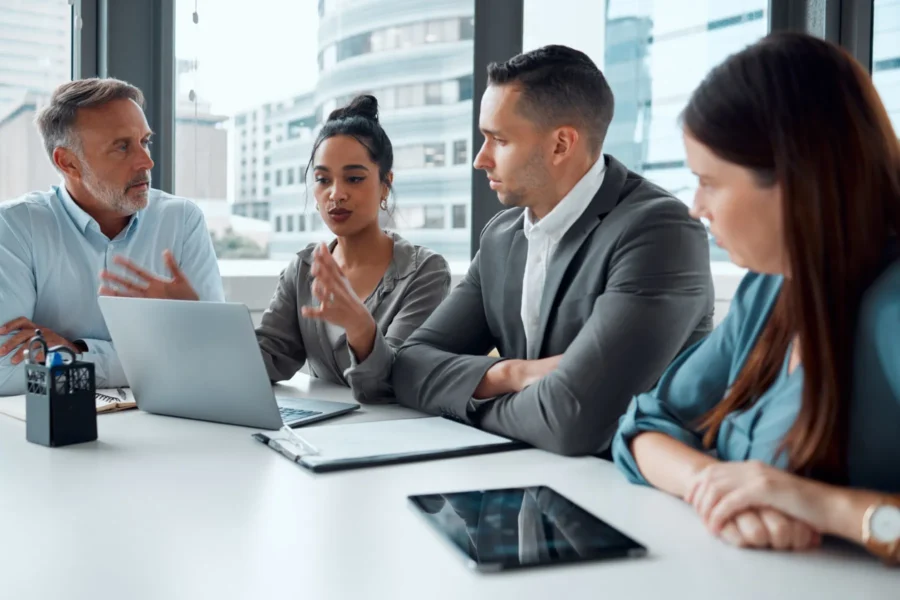 Four people sit at a table in a modern office, having a discussion with a laptop, documents, and a tablet in front of them.