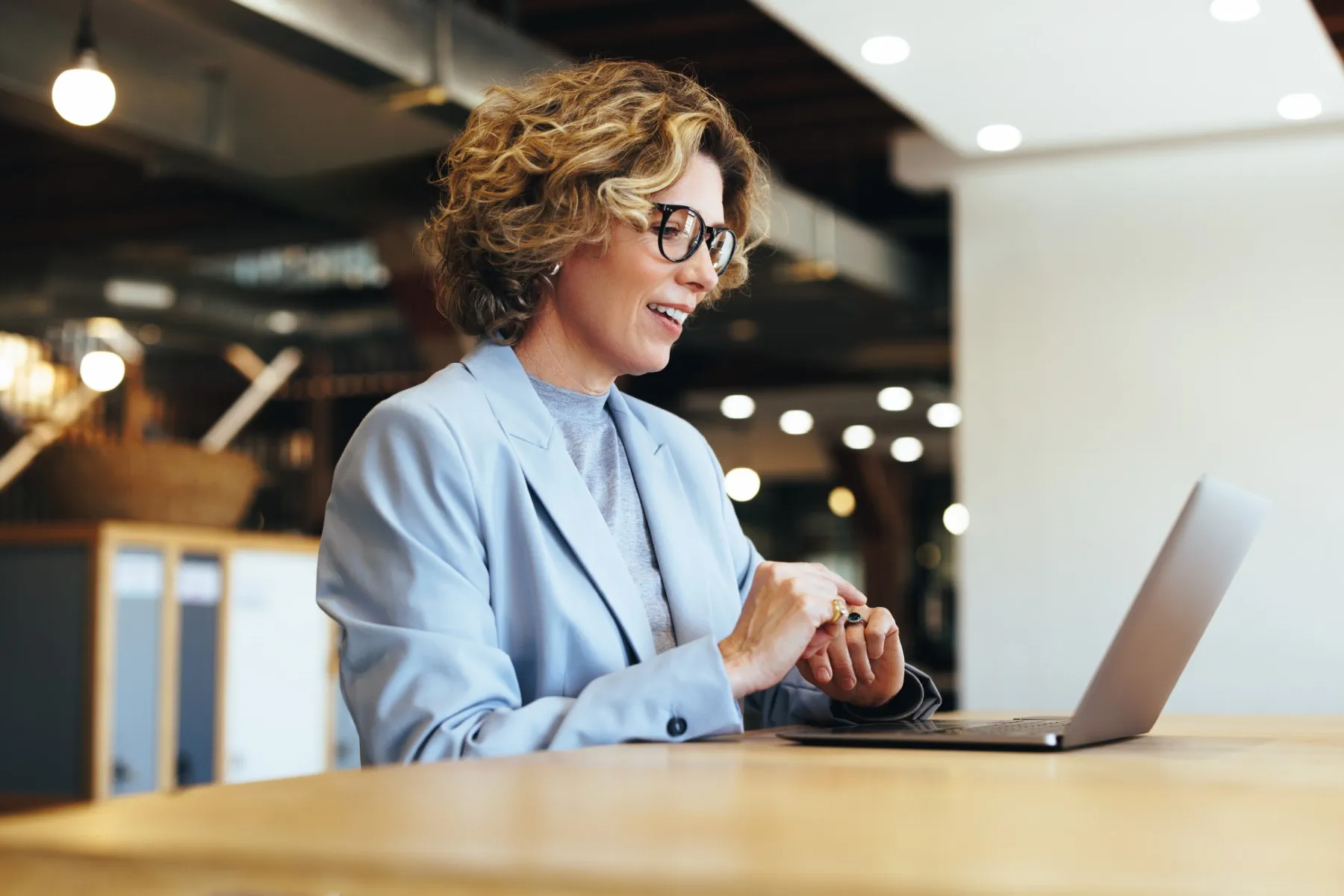 A woman wearing glasses and a light blue blazer sits at a desk, smiling and looking at a laptop in a modern office setting.