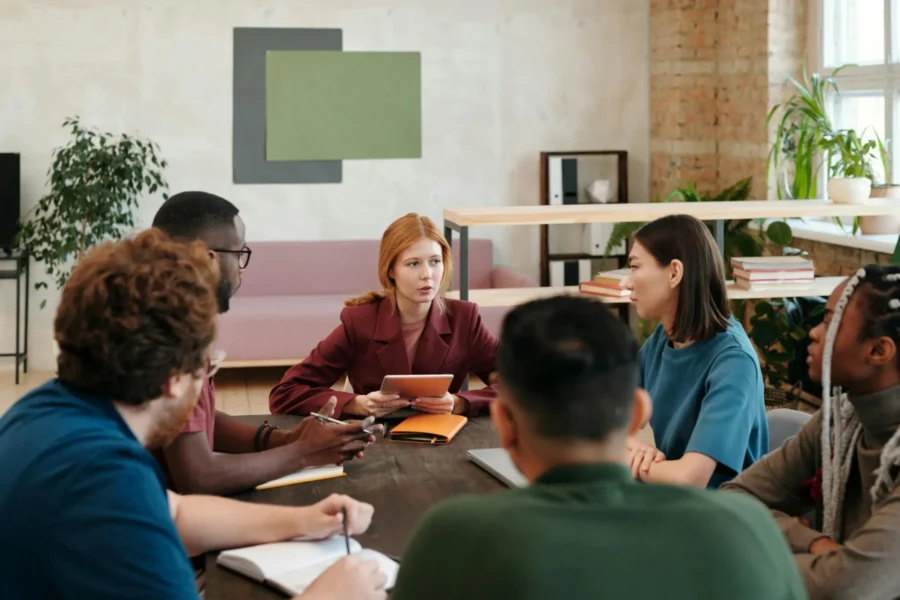 Six people sit around a conference table having a discussion, with one person holding a tablet and leading the meeting.