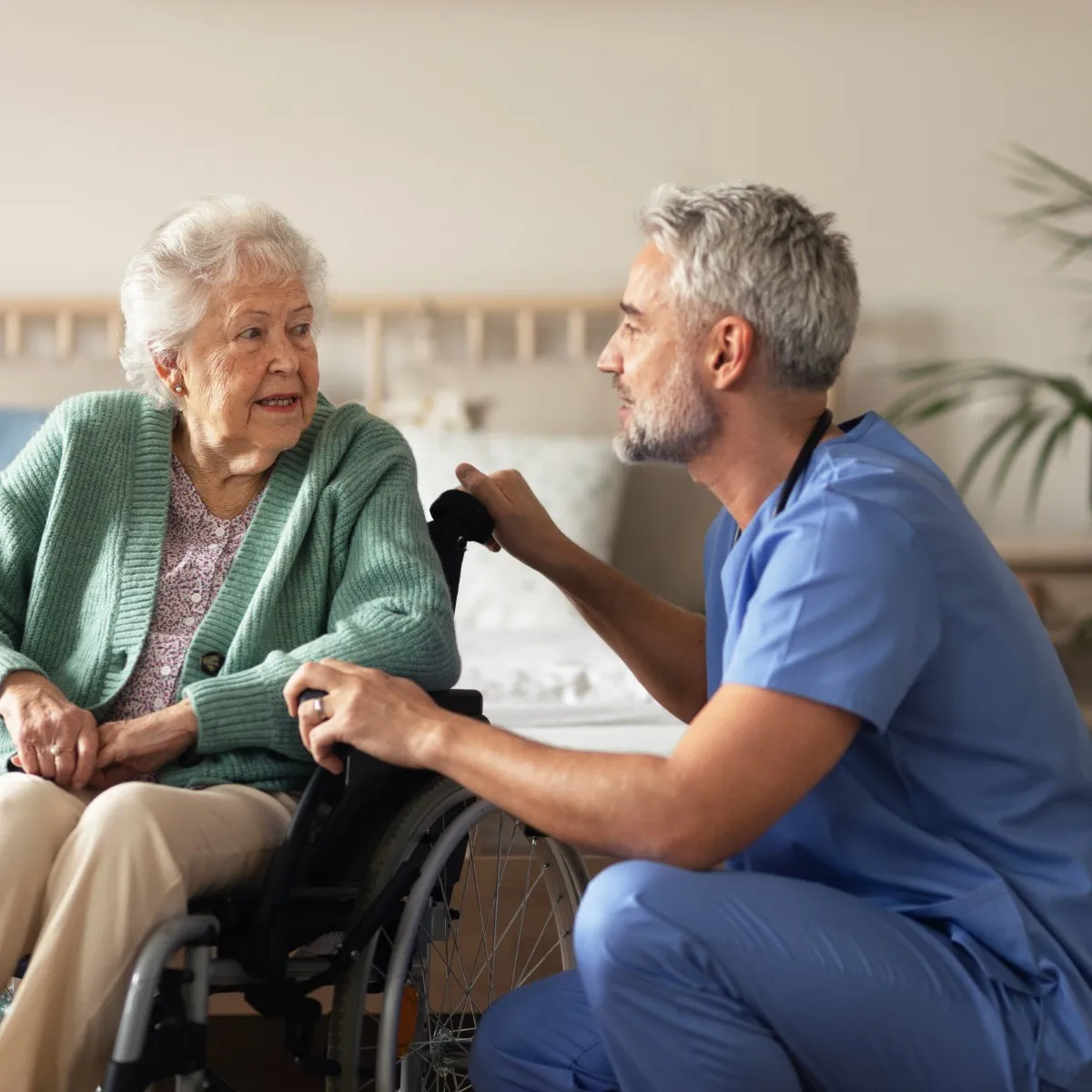 An elderly woman in a wheelchair talks with a male nurse in blue scrubs, who is kneeling beside her in a well-lit room.