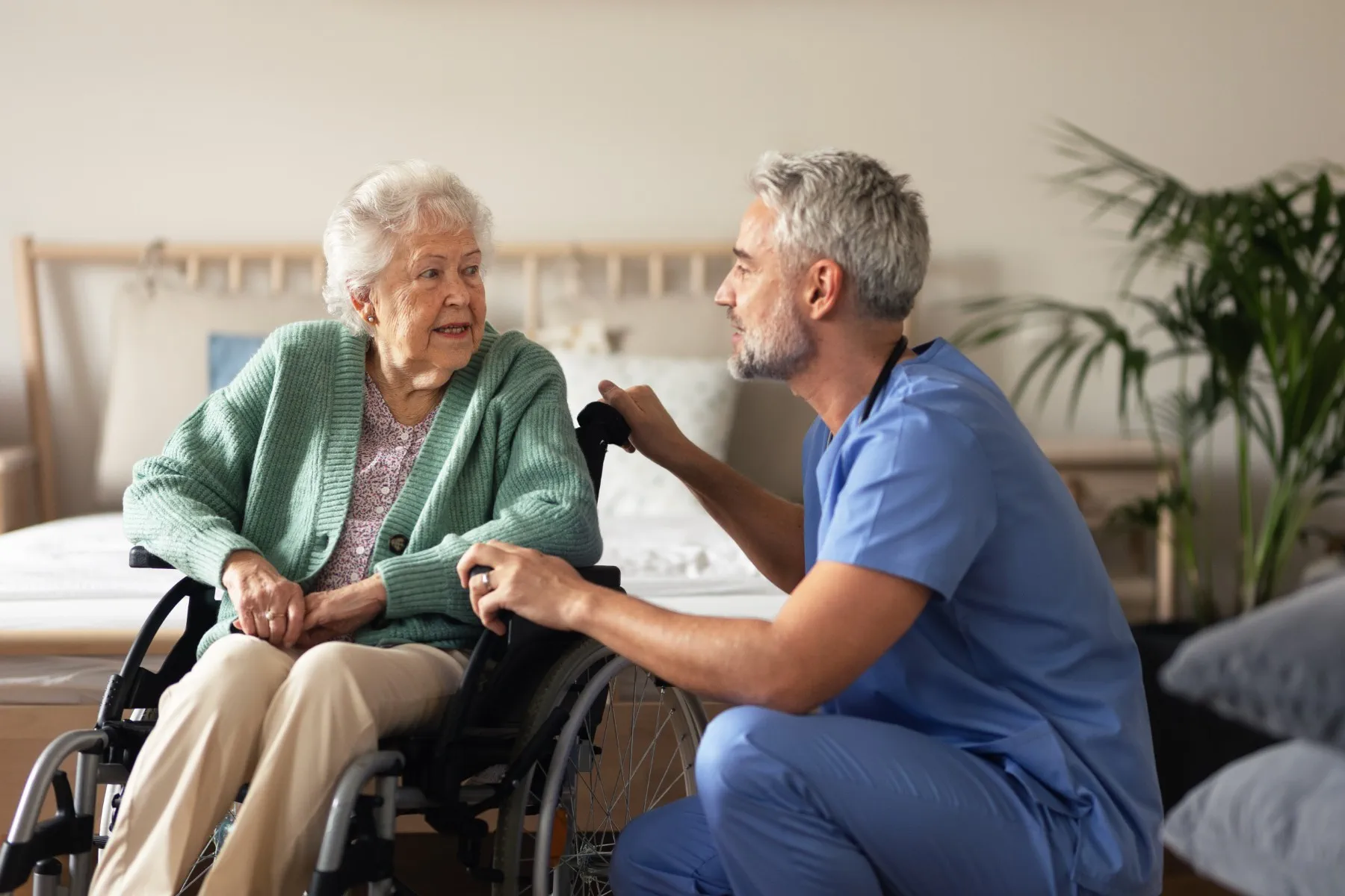 A healthcare professional in blue scrubs talks to an elderly woman in a wheelchair in a bedroom setting.