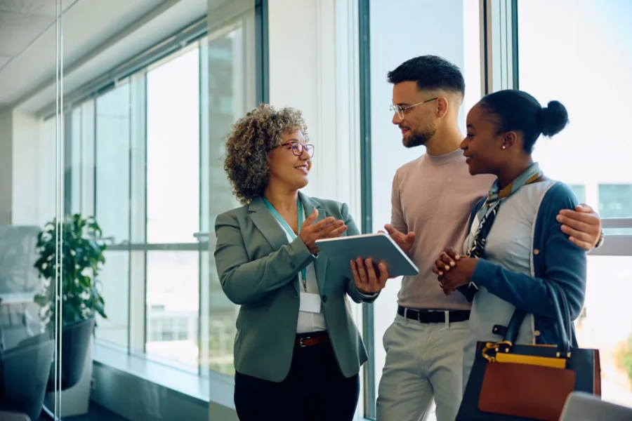 Three people stand indoors near large windows, conversing and smiling. One person holds a tablet, and all appear to be engaged in a professional discussion.