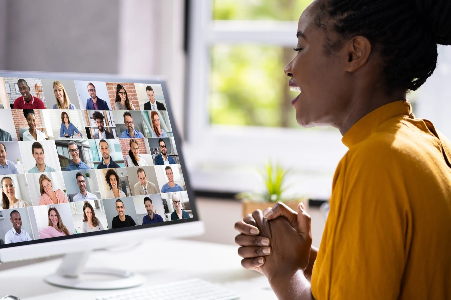 A woman in a yellow top participates in a video conference call with multiple people shown on her computer screen.