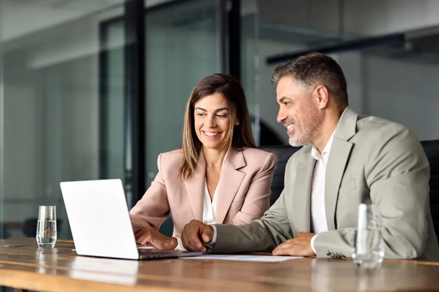 Two people in business attire sit at a desk, smiling and looking at a laptop screen, with papers and glasses of water in front of them in a modern office setting.
