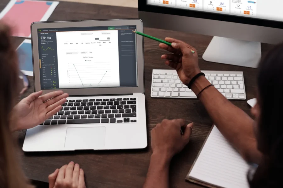 Two people review data charts on a laptop screen, one pointing with a pencil, while a desktop computer, keyboard, and notebook are on the desk.