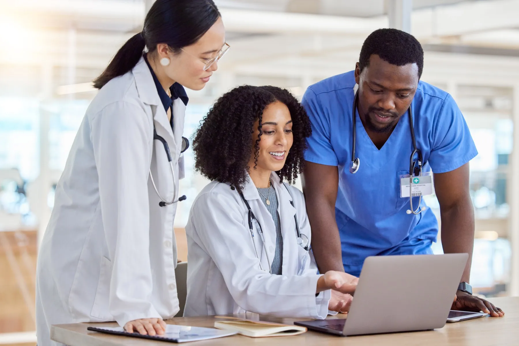 Three medical professionals in white coats and scrubs discuss information displayed on a laptop in a clinical setting.