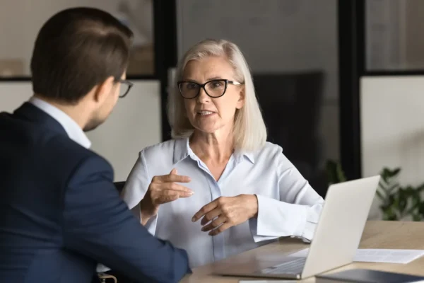 Two people in business attire sit at a desk with a laptop, engaged in a serious conversation in an office setting.