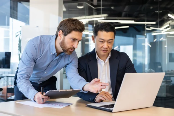 Two men in business attire are working together at a desk, with one pointing at a laptop screen and the other holding a clipboard in an office setting.