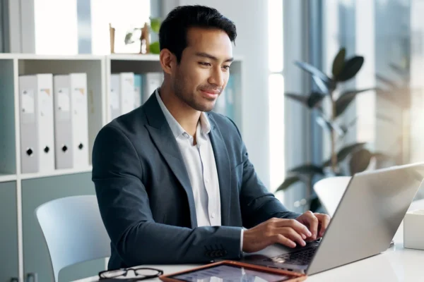 A man in a suit sits at a desk typing on a laptop in a modern office with shelves and plants in the background.