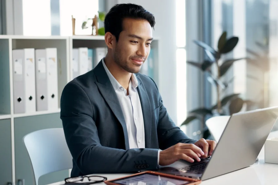 A man in a suit sits at a desk typing on a laptop in a modern office with shelves and plants in the background.