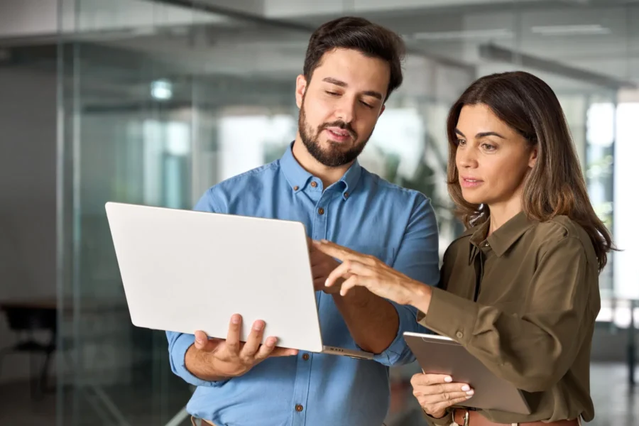 Two people standing indoors, looking at a laptop screen together; one person holds the laptop while the other points at the screen and holds a tablet.