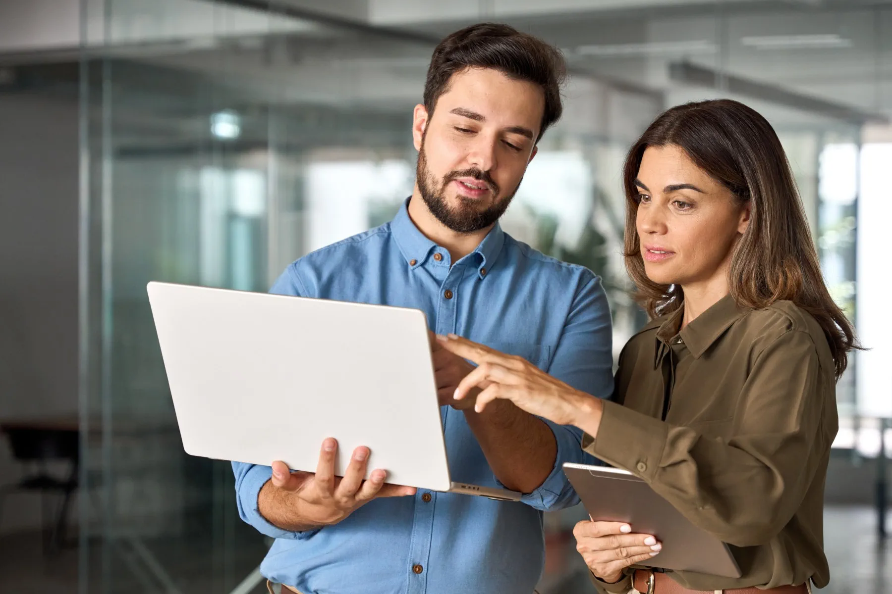 Two people standing indoors, looking at a laptop screen together; one person holds the laptop while the other points at the screen and holds a tablet.