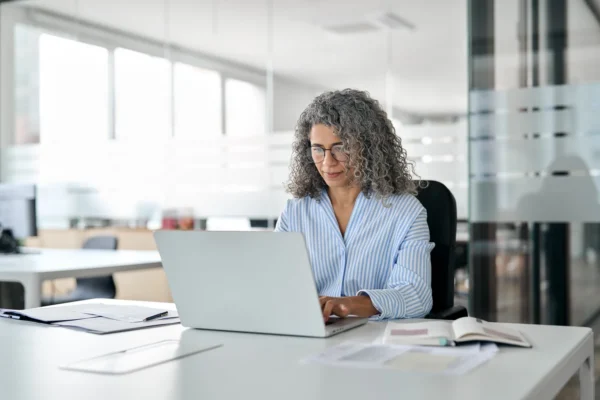 A woman with curly gray hair sits at a desk in a modern office, working on a laptop with documents and an open notebook nearby.