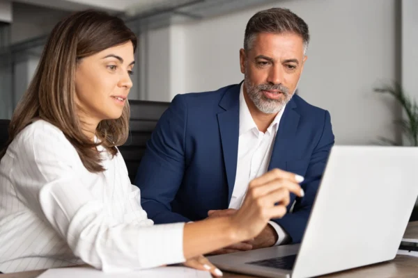 Two business professionals sit at a desk looking at a laptop screen, with one person pointing at the screen while discussing something.