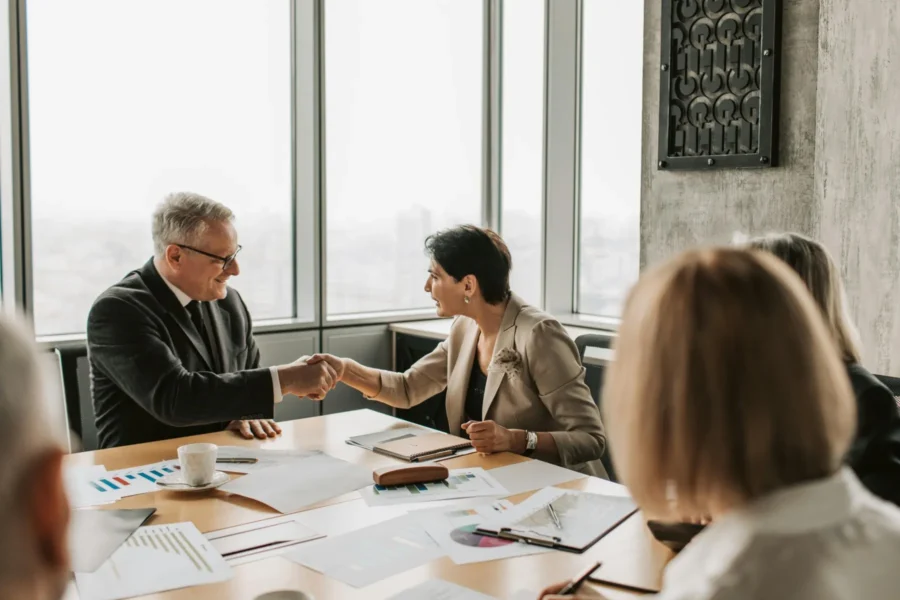 Two businesspeople shake hands at a conference table during a meeting, with documents, charts, and coffee cups on the table.