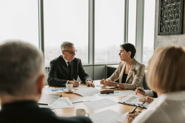 Four people in business attire sit around a conference table with documents and charts, engaged in a meeting in a modern office with large windows.