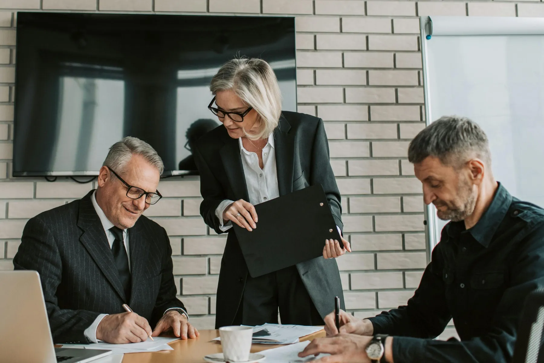 Three business professionals in suits review documents at a conference table, with a laptop, papers, and a coffee cup in front of them, in a modern office setting.