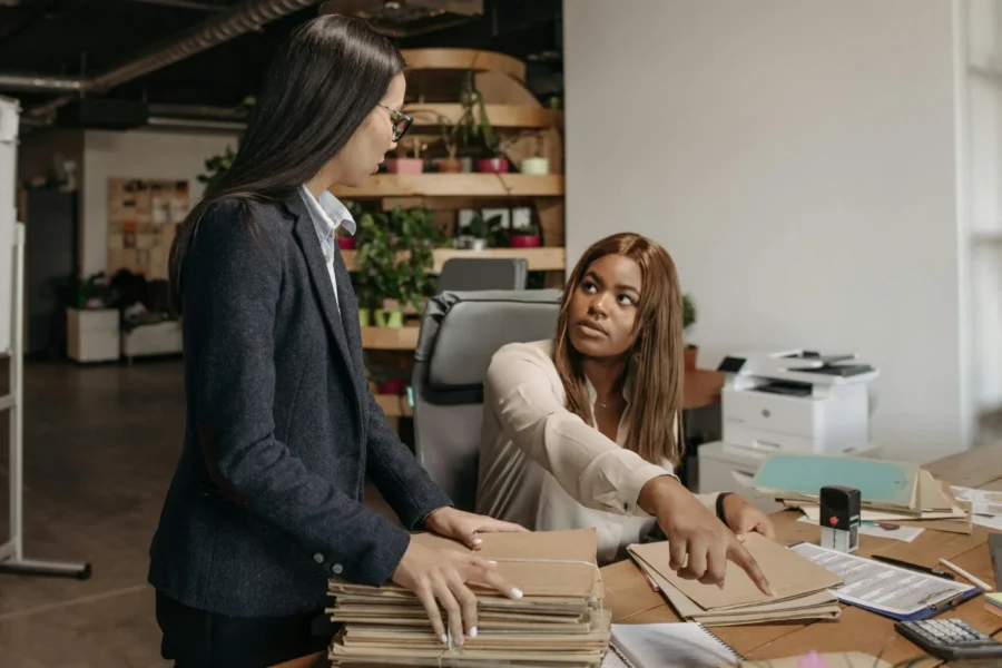 Two women in an office discuss paperwork at a desk covered with files, documents, and office supplies. One is seated and pointing, while the other stands holding folders.