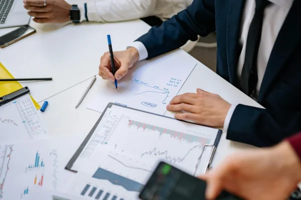 Person in business attire draws a line graph on paper surrounded by printed financial charts and documents during a meeting.