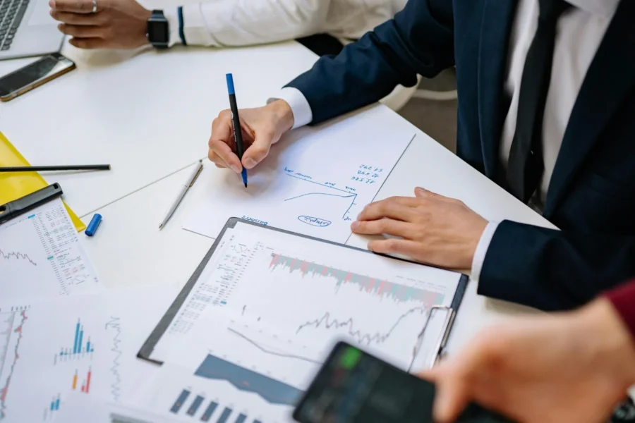 Person in business attire draws a line graph on paper surrounded by printed financial charts and documents during a meeting.