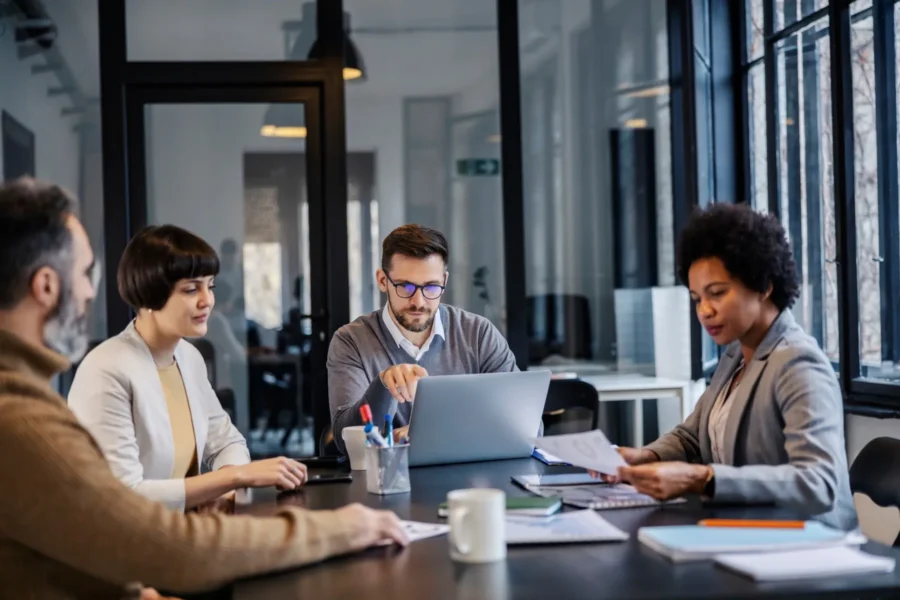 Four people sit around a conference table in a modern office, engaged in discussion with documents and a laptop on the table.