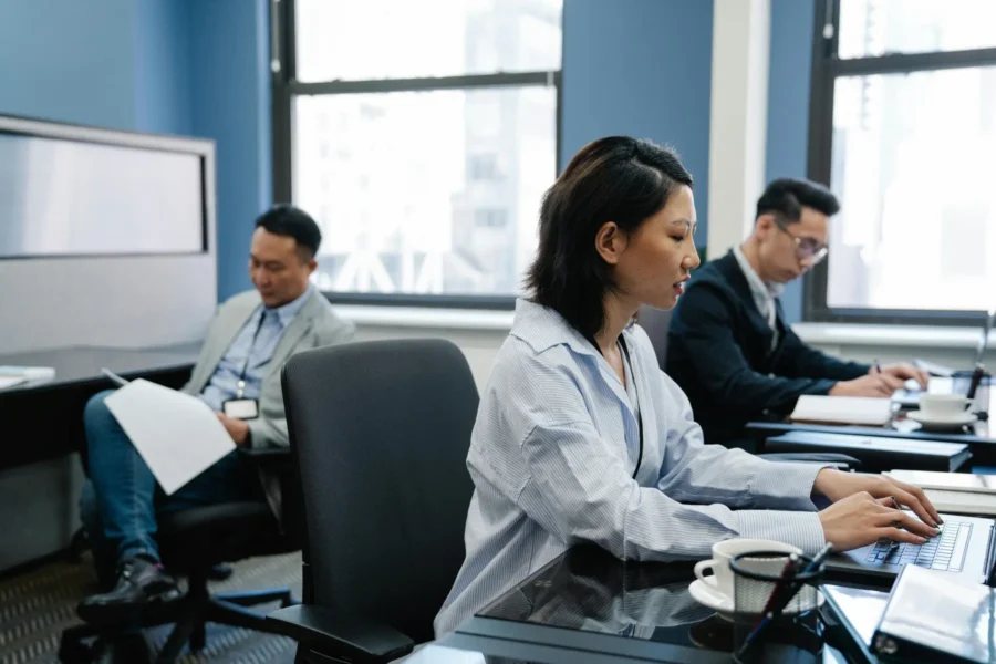 Three people working in a modern office; two are using laptops at desks while one reads documents in the background. Coffee cups and office equipment are visible.