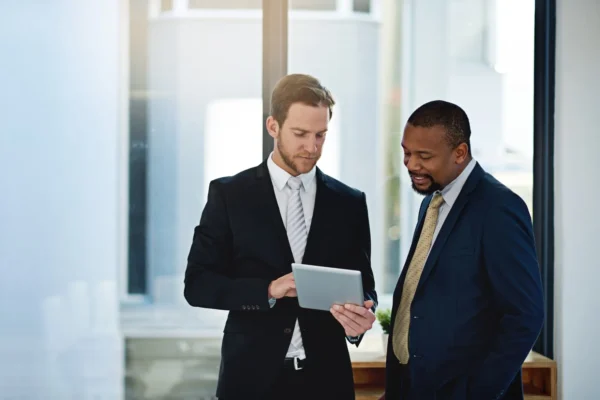 Two men in business suits stand by a window in an office, looking at a tablet device together.