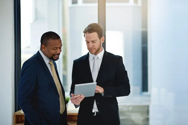 Two men in business suits stand indoors by a window, looking at a tablet device together.