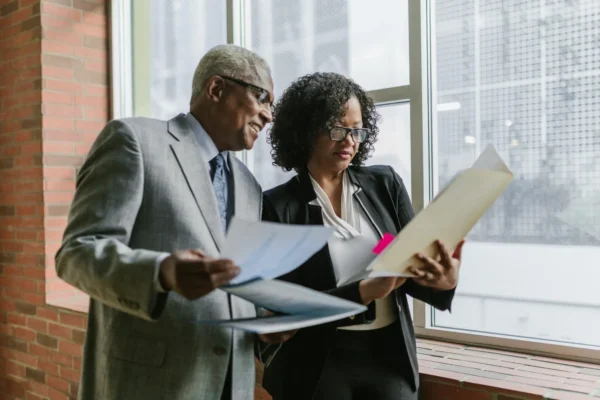 Two business professionals review documents together by a large window in an office setting.