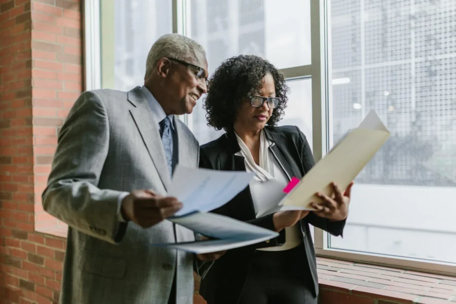 Two business professionals review documents together by a large window in an office setting.