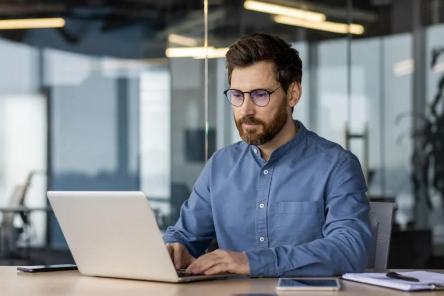 A man with glasses and a beard works on a laptop at a desk in a modern office setting, with papers and a smartphone nearby.