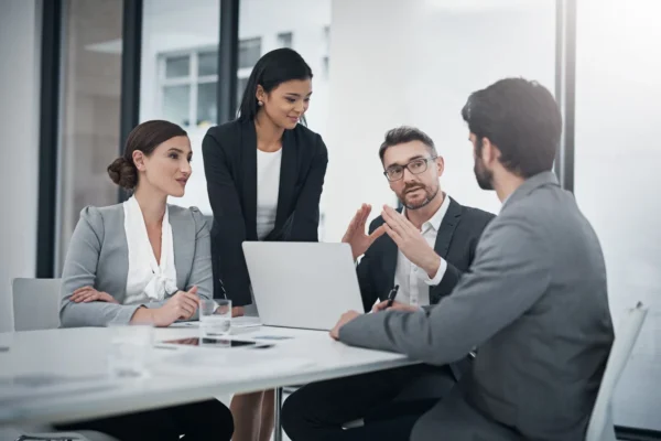 Four business professionals in formal attire have a discussion around a table with a laptop and documents in an office setting.