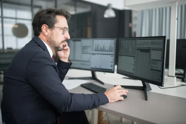 Man in a suit sits at a desk using a mouse and looking at financial data and charts on two computer monitors in an office.