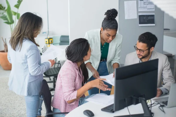 Four colleagues discuss documents around a desk with a computer in a modern office setting. Two are standing while two sit, reviewing paperwork together.