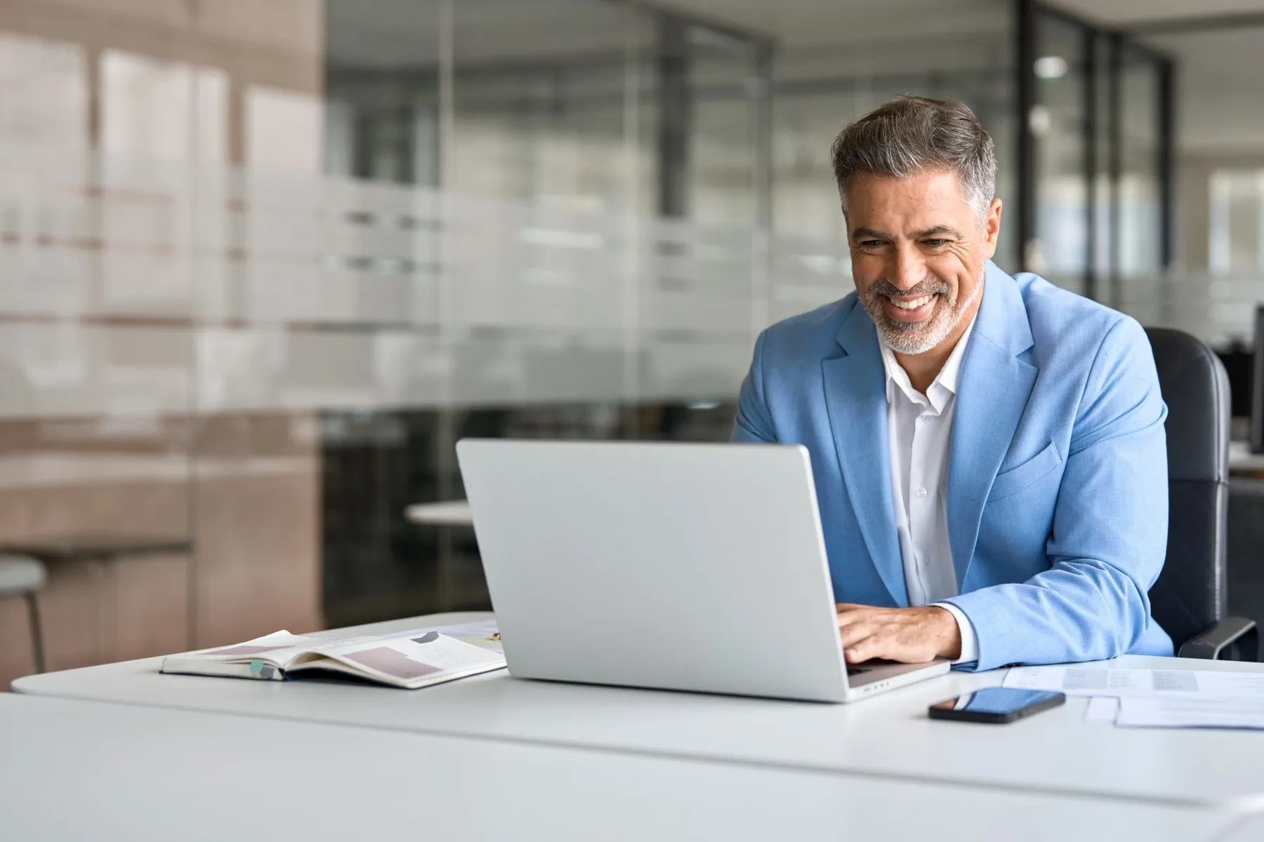 Smiling man in a light blue blazer works on a laptop at a desk in a modern office, with a notebook, documents, and phone nearby.