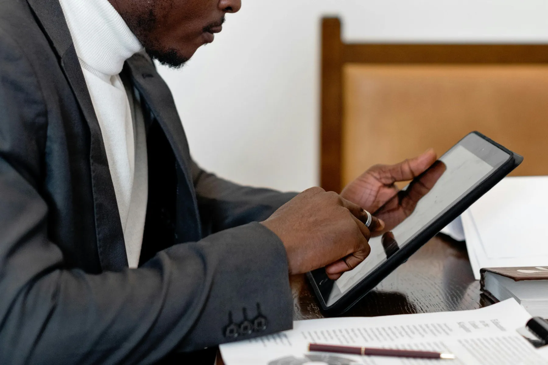 A person in a suit uses a tablet at a desk with papers, a pen, and a book.