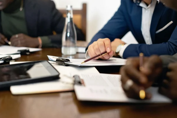 Several people in business attire sit at a table with documents, clipboards, a tablet, and a glass water bottle, writing and discussing paperwork.