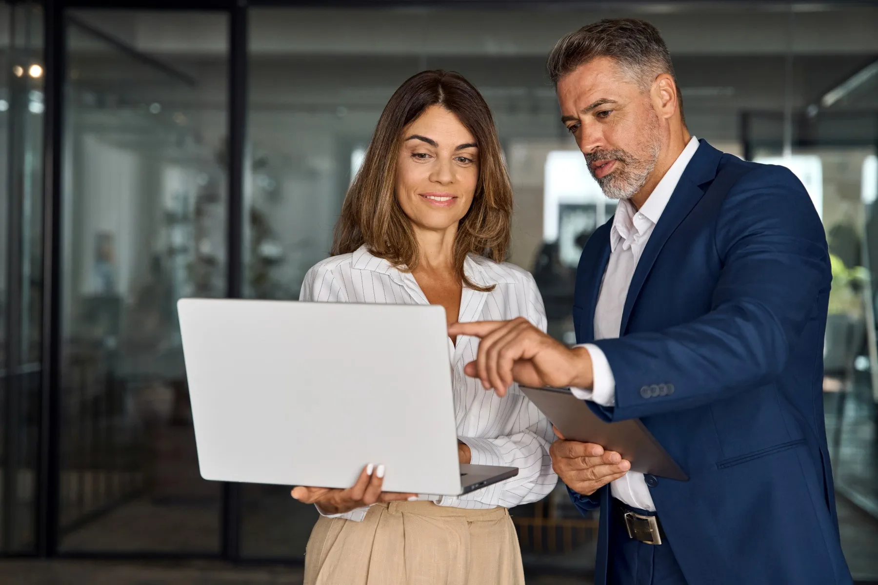 Two business professionals stand together in an office, looking at a laptop screen while one points at it, both appearing focused and engaged.