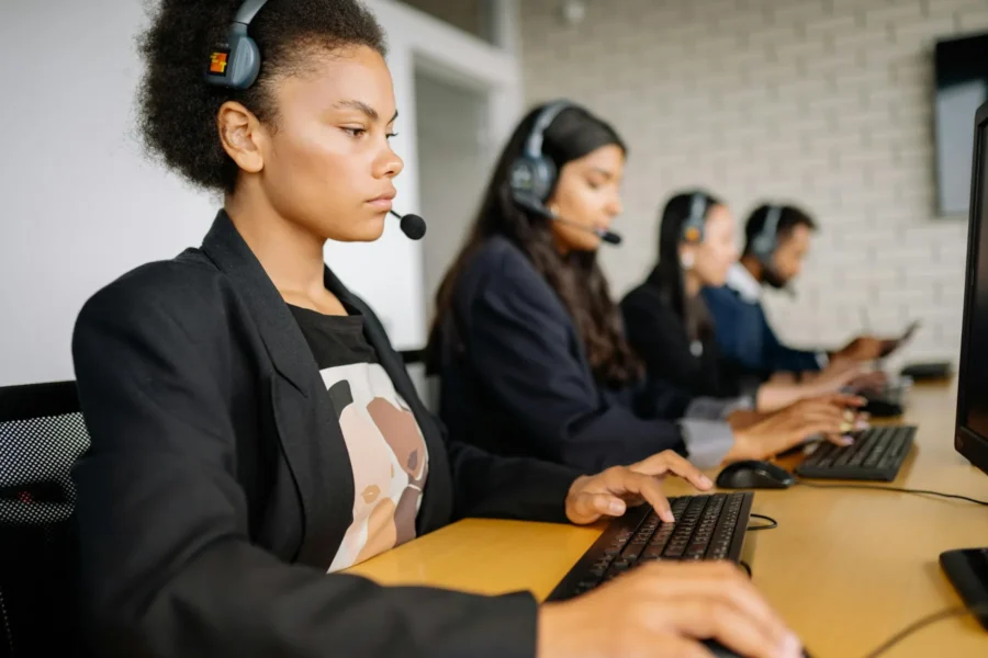 Four people wearing headsets sit in a row at desks, typing on keyboards and looking at computer monitors in an office setting.