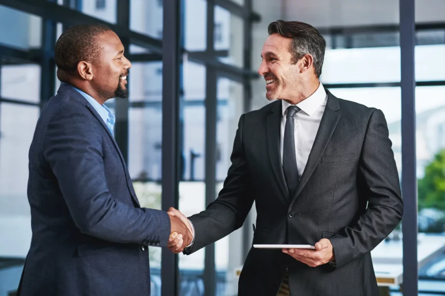Two men in business attire shake hands and smile at each other in a modern office setting, with one holding a tablet.
