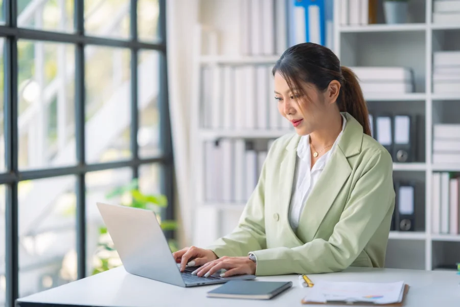 A woman in a light green blazer works on a laptop at a desk in a modern office with shelves and large windows in the background.