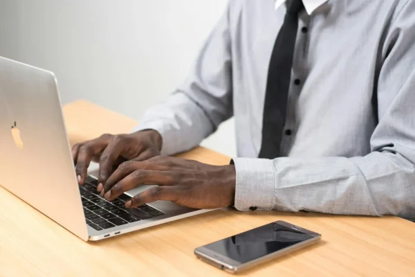 A person in a gray shirt and black tie types on a laptop at a wooden desk with a smartphone placed beside the computer.