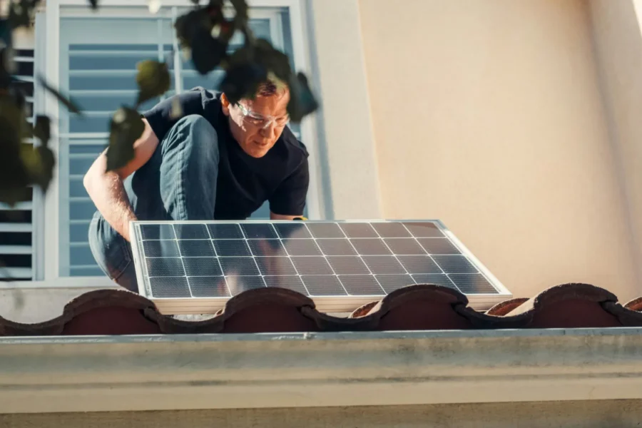 A person wearing safety glasses installs a solar panel on the roof of a beige building.
