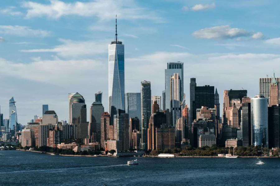 Skyline view of Lower Manhattan with skyscrapers, including One World Trade Center, seen from across the Hudson River under a partly cloudy sky.