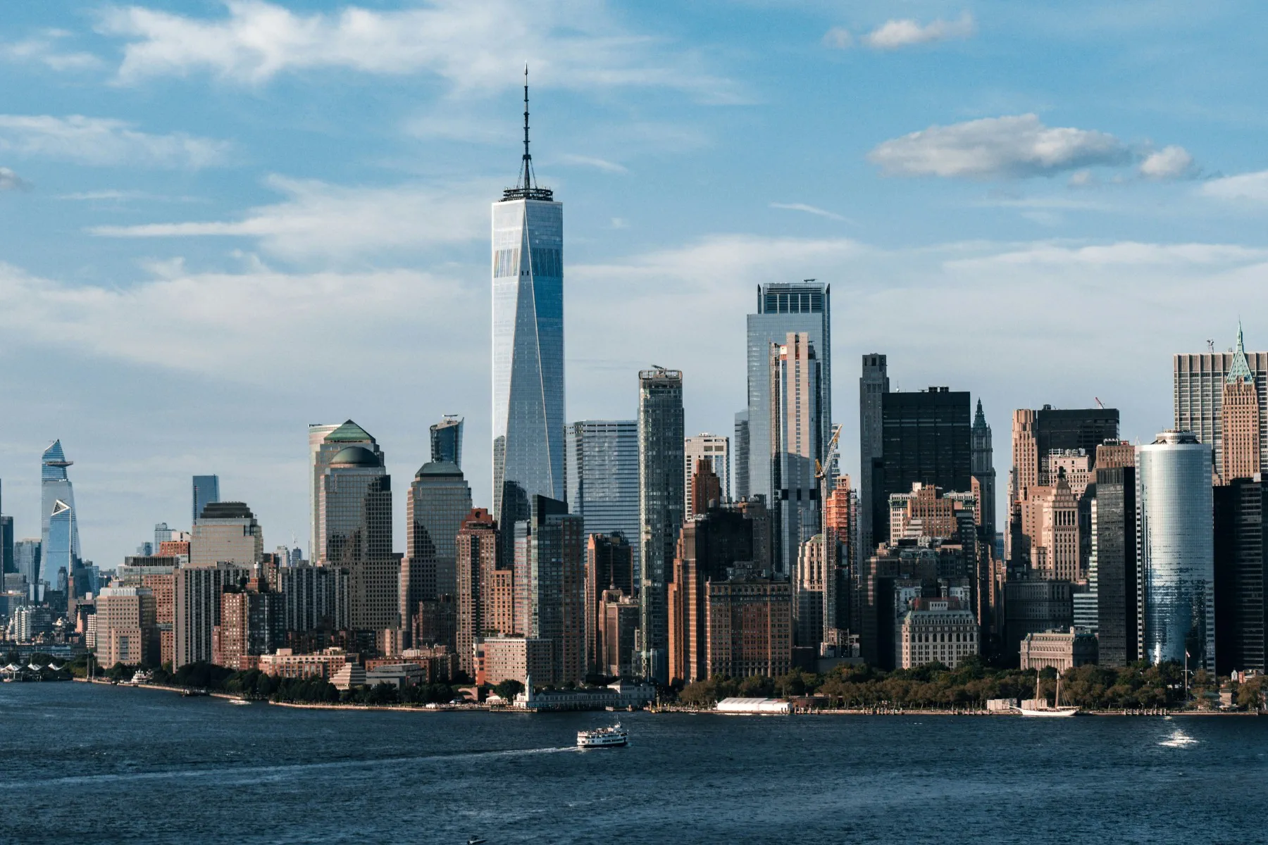Skyline view of Lower Manhattan with skyscrapers, including One World Trade Center, seen from across the Hudson River under a partly cloudy sky.