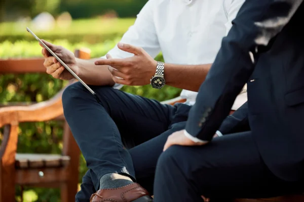 Two people in business attire sit on a bench outdoors, one holding a tablet and gesturing while the other listens.