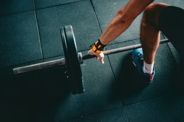 A person wearing blue shoes and a wrist strap grips a barbell with weight plates on a gym floor.