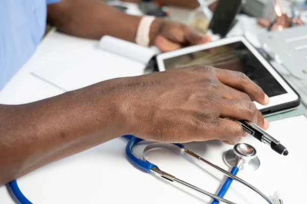 A person’s hand holding a pen rests on a tablet, with a stethoscope and notebooks on a white desk, suggesting a medical work environment.
