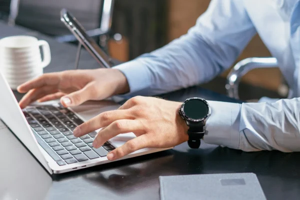 Person in a light blue shirt typing on a laptop at a desk, with a smartwatch on their wrist, a notebook, a smartphone, and a coffee cup nearby.