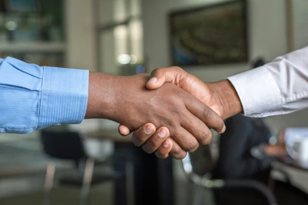 Two people wearing dress shirts shake hands in an office setting, with blurred furniture and a person in the background.